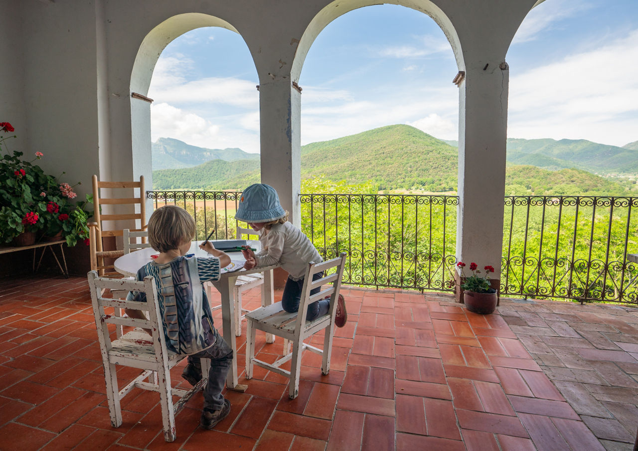 Dos infants dibuixant en una taula ubicada a la primera planta de la Masia Mas Garganta amb vista a la Vall d'en Bas.