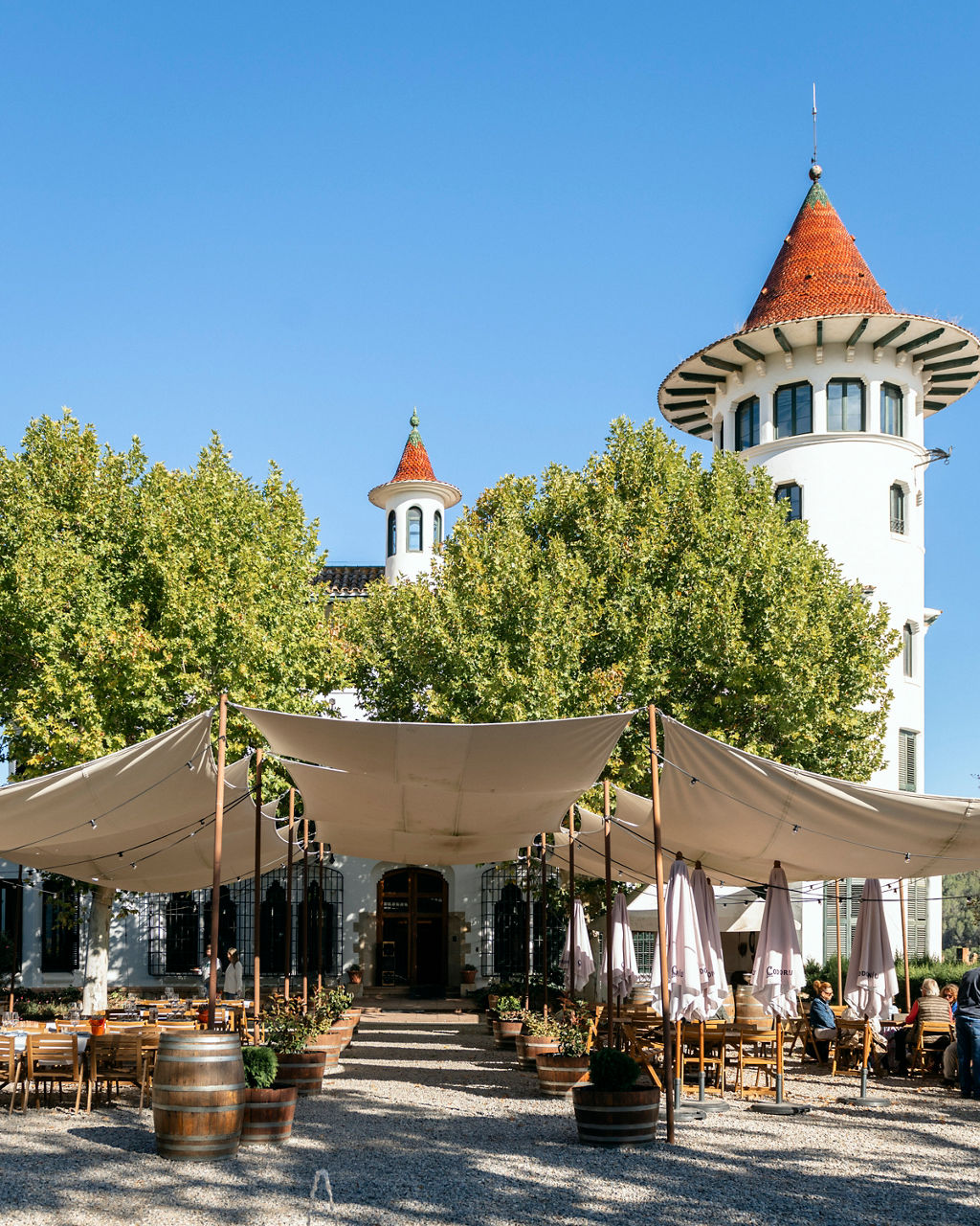 Terrassa del restaurant La Torre de Codorníu, ubicat a Sant Sadurní d’Anoia, amb tendals de tela i taules i cadires de fusta, amb alguns comensals asseguts en un dia assolellat.