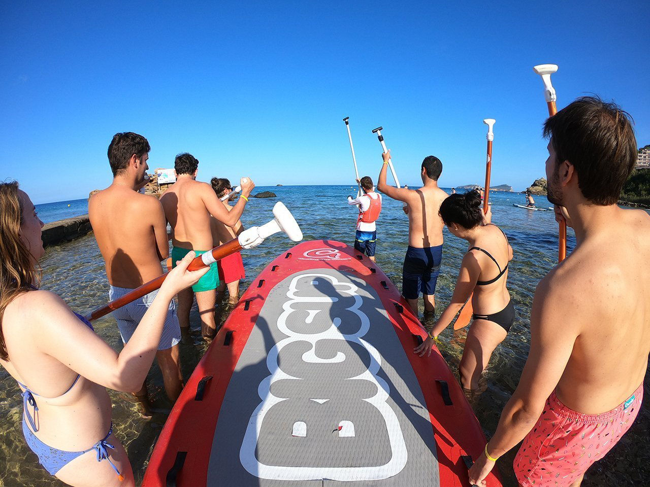 Participants de l’activitat de 'team building' "Blue Beach Sports", organitzada per l’empresa High Fidelity Collective, entrant a l’aigua des d’una platja, a punt per practicar Big SUP, una modalitat de paddle surf que consisteix a remar sincronitzats sobre una taula gegant.