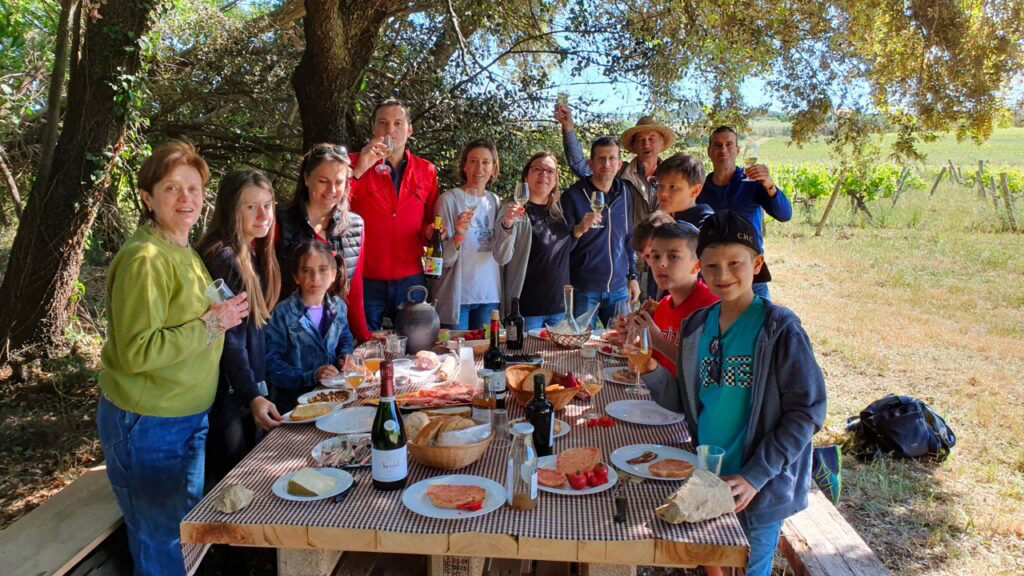 Un grup de persones reunides al voltant d'una taula de pícnic parada, amb diversos aliments i begudes com embotit, oli, vi i sucs; situada en una zona d'ombra amb vista a les vinyes de Can Suriol.