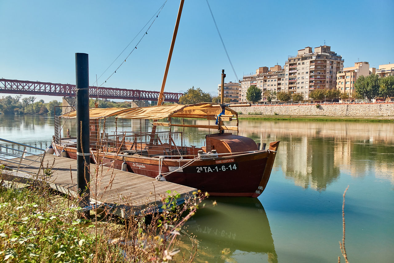 Embarcació típica anomenada "Lo Sirgador", amarrada al riu Ebre al seu pas per Tortosa. Al fons es pot veure l'antic pont del ferrocarril (construït entre 1865 i 1868), avui convertit en una passarel·la peatonal que forma part de l'itinerari de la Via Verda.