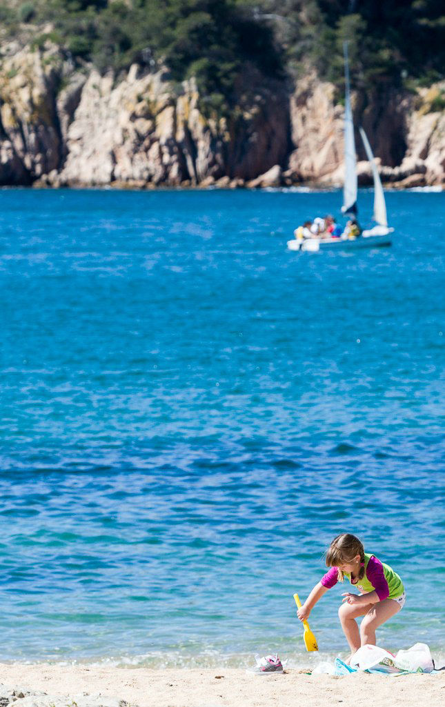 Nena jugant amb la sorra a la platja de Sant Feliu de Guíxols amb un veler al fons.