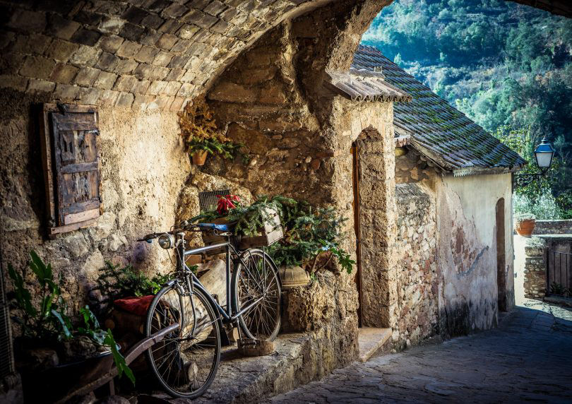 Grand Tour de Catalunya. Tram 1. Carrer empedrat, amb plantes i un arc de pedra, a la població de Mura. Recolzada en una paret hi ha una bicicleta decorada que fa de jardinera.